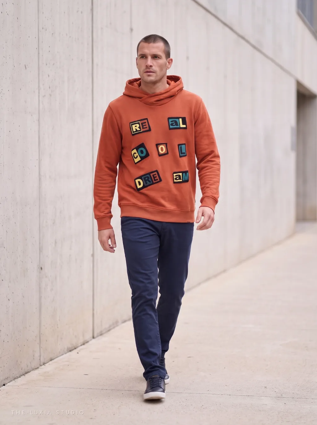 Después, ambiente: modelo creada con IA con la misma sudadera naranja con parches caminando por un exterior de hormigón